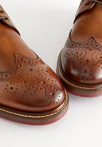 Paire de chaussures brogues en cuir marron avec perforations décoratives et semelles épaisses cousues, sur un fond clair uni.