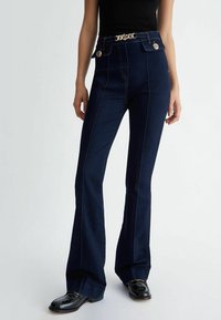 Tmavě modré široké džíny s akcenty zlatých knoflíků a dekorativní zlatou sponou na pásku. Vyznačují se kontrastním stehováním a hladkou denímovou texturou.