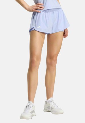 Femme portant un short de sport bleu clair et des chaussures de course blanches, debout avec une main sur la hanche sur un fond uni.