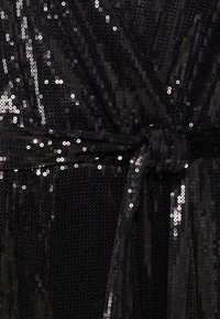 Zwarte paillettenjurk met een gestructureerd oppervlak, voorzien van een strikdetail in de taille. Reflecterende pailletten creëren een glinsterend effect door de hele jurk.