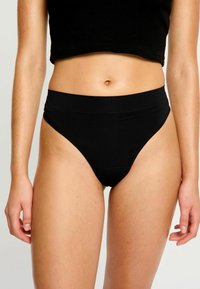 Femme portant des sous-vêtements noirs à coupe haute et un haut noir court, montrant le ventre et les bras devant un fond clair.