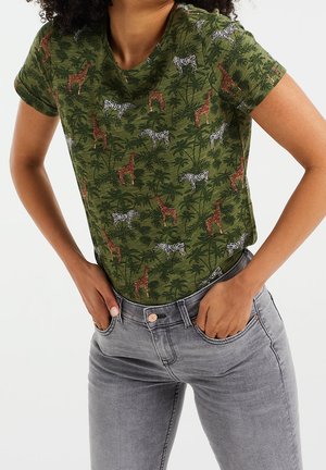 Persoon draagt een groen shirt met korte mouwen met giraffe- en zebrapatroon en lichtgrijze jeans, handen rustend in de voorzakken.