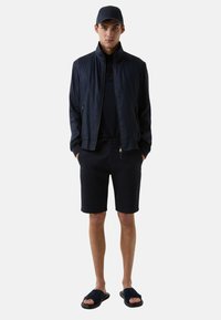 Veste aviateur marine, polo assorti, shorts et tongs. Tissu doux, poches zippées et design ajusté, le tout dans des teintes sombres.