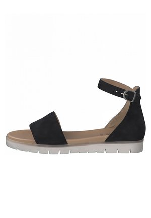 Sandale plate noire avec boucle de cheville et large sangle avant, semelle intérieure beige et semelle en caoutchouc blanc avec des nervures.