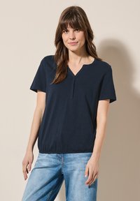 Mörkblå t-shirt med kort ärm och V-ringning, avslappnad passform och elastisk nederkant; slät tyg med en lätt lyster. Bärs med ljusblå jeans.