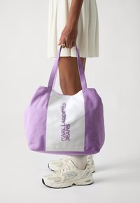 Tygväska med lavendelfärgad tyg, en vit mittpanel och texten "KARL LAGERFELD JEANS" i lila. Dubbelhandtag för bärande.