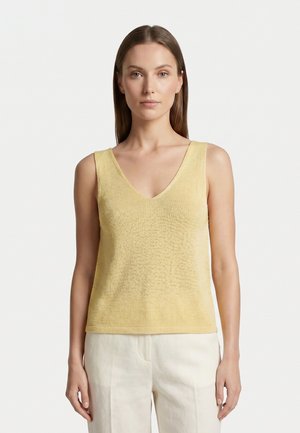 Donna con capelli castani lisci che indossa un top giallo pallido senza maniche con scollatura a V e pantaloni beige chiaro, in piedi davanti a uno sfondo bianco semplice.