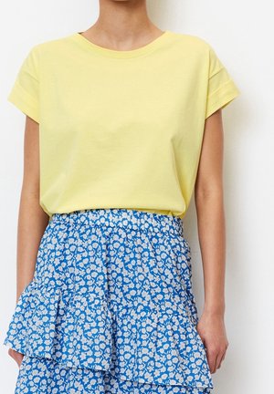Person trägt ein gelbes Kurzarmshirt, das in einen blauen Rock mit weißen Blumenmustern und gerafften Stufen gesteckt ist.