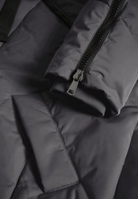 Isolatiejas van donkergrijs nylon, met een ritssluiting aan de voorkant, ribgebreide manchetten en een gladde, gewatteerde textuur met een matte afwerking.