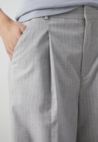 Main reposant dans la poche d'un pantalon gris clair à fines rayures avec un pli à l'avant, assorti à un haut gris clair côtelé.