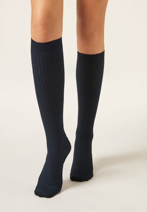 Gambe che indossano calze nere a coste fino al ginocchio, in piedi su una superficie chiara con uno sfondo neutro.