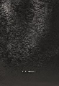 Black textured leather surface with fine grain and small silver metal "COCCINELLE" brand logo centered near the bottom.