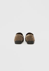 Paire de mocassins en suède marron avec semelles noires vus de dos sur un fond clair uni.