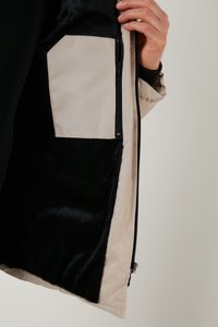 Fodera in pelliccia nera con un soprattuto in tessuto beige; presenta una tasca con zip. La giacca ha una texture liscia e un design strutturato.