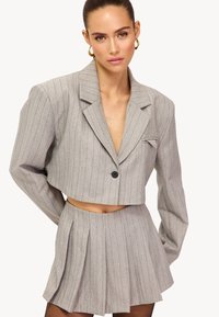 Blazer court gris à fines rayures avec larges revers et un seul bouton, associé à une jupe plissée avec une découpe à la taille.
