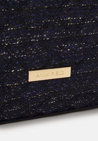 Zwarte gevlochten handtas met blauwe en witte draden, met een gouden metalen logoplaatje waarop "ANNA FIELD" staat. Gestructureerd oppervlak met een gewatteerd patroon.
