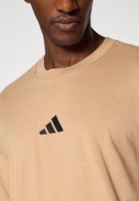 T-shirt en coton beige à manches courtes, arborant un logo Adidas noir brodé sur le côté gauche de la poitrine. Texture douce et lisse.