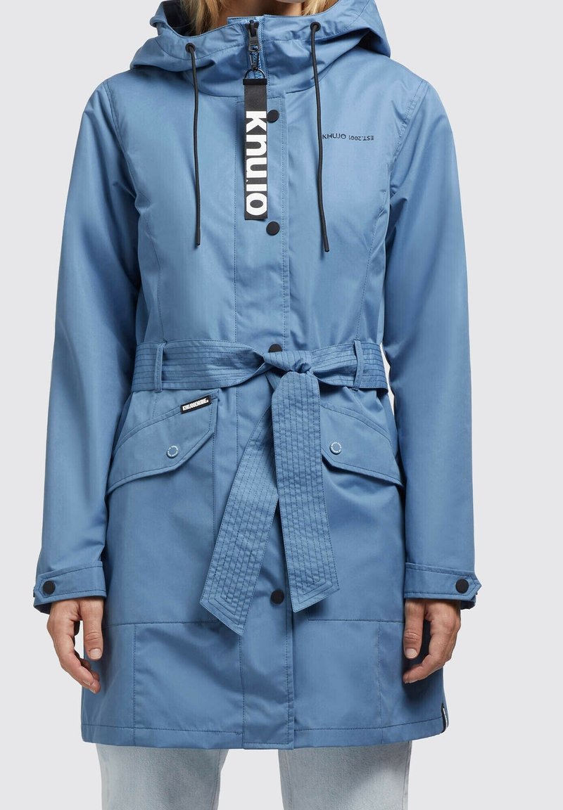 Ljusblå vattentät jacka med huva, justerbara dragsnören, skärp och knappfickor. Har logotyp och vadderad skärpdetalj.