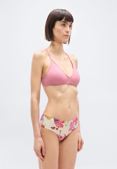 Femme portant un haut de bikini triangle rose et un bas de bikini taille haute à motif floral, debout devant un fond uni clair.