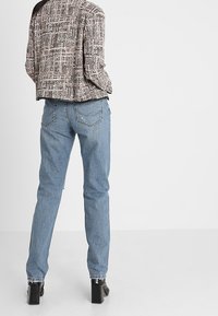 Szara teksturowana krótka kurtka w zestawieniu z jasnoniebieskimi prostymi dżinsami i czarnymi botkami na słupku, pokazana z tyłu.