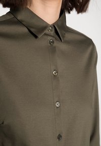 Camisa de botones verde oliva con cuello clásico, que presenta una textura sutil y botones metálicos, cortada de manera ajustada por encima del área del pecho.