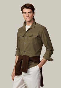 Camisa de color verde oliva con botones y dos bolsillos en el pecho, mangas remangadas, y un suéter de punto marrón oscuro atado alrededor de la cintura; pantalones color marfil.