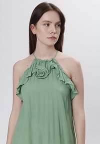 Top verde senza maniche con dettagli arricciati e un particolare floreale sul colletto. Realizzato in tessuto leggero con una texture morbida.