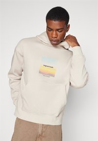 Beige hoodie met een voorzak en een rechthoekige grafiek met "Originals Studio" in een gradient van pastelkleuren. Zachte textuur.