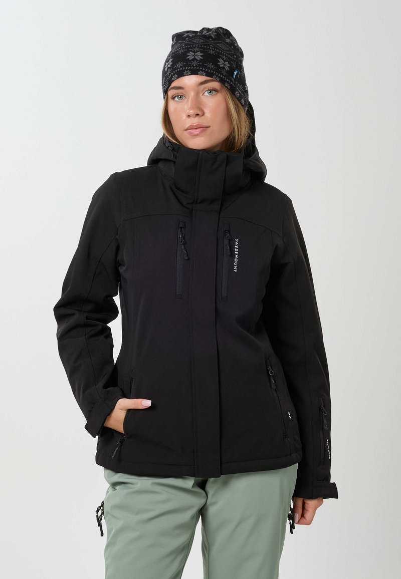 Vrouw draagt een zwarte winterjas, een donkere gebreide muts met patroon en lichtgroene broek, handen in de zakken, gericht naar voren op een effen achtergrond.
