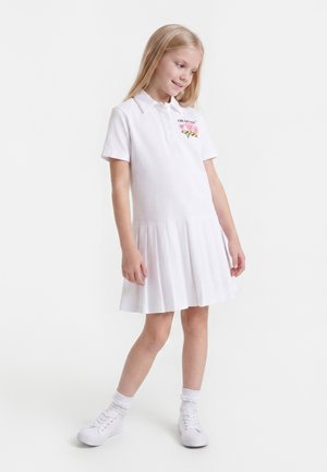 Jeune fille debout portant une robe polo blanche à manches courtes avec broderie florale, des chaussettes blanches et des baskets blanches sur fond blanc.