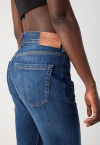 Personne portant un jean bleu taille haute avec une étiquette en cuir marron Marc O’Polo, visible du milieu du dos jusqu’au milieu de la cuisse sur un fond uni.