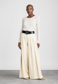 Fehér hosszú ujjú felső, krémszínű, bőszárú, pliszírozott nadrággal és fekete övvel párosítva. Lapos, átszőtt mintás szandálok kivágásokkal.