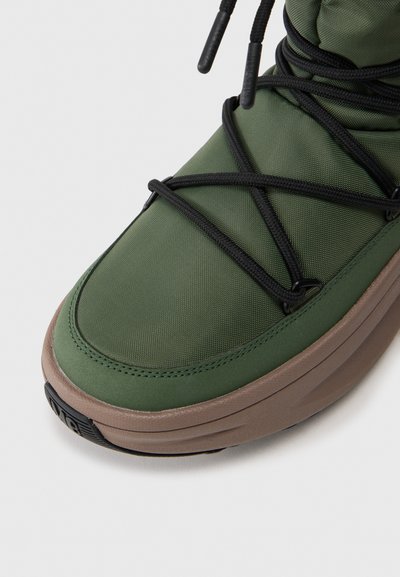 Zapatilla verde con un upper liso y texturizado, cordones negros y una suela gruesa marrón. Diseñada con una punta redondeada y costuras mínimas.