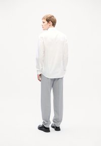 Chemise blanche à manches longues avec un col pointu, associée à un pantalon gris clair et des chaussures noires, vue de dos.