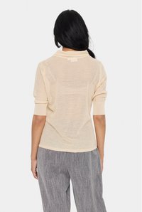 Maglione lavorato a maglia color pesca chiaro con maniche corte, texture a trama aperta e colletto rotondo. Indossato con pantaloni grigi a gamba larga.