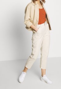 Beżowa krótka kurtka, rdzawy top na guziki oraz białe spodnie z wysokim stanem, zestawione z białymi sneakersami. Gładka tkanina i luźny krój.