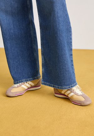 Osoba w szerokich niebieskich dżinsach i beżowych sneakersach z białymi paskami stojąca na podłodze w kolorze musztardowym.