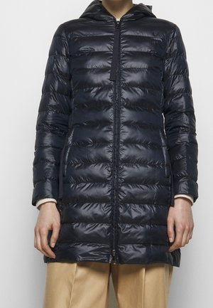 Osoba nosząca długą czarną pikowaną kurtkę puchową z kapturem, zapinaną na zamek, oraz beżowe spodnie widoczne od połowy uda w dół.