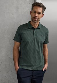 Lyhyttukkainen ruskeahiuksinen ja parran sänkeä oleva mies pukeutunut tummanvihreään kuvioituun poolopaitaan ja tummansinisiin housuihin seisoo betoniseinää ja valkoista seinää vasten.