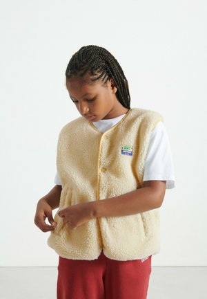 Beige fleecevest met een zachte textuur, afgeronde zoom en gele bies. Voorzien van een logopatch en knopen aan de voorkant. Gedragen over een wit shirt.