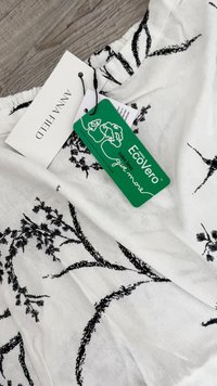 Tela blanca con estampado botánico negro, que presenta dos etiquetas: una etiqueta blanca de "Anna Field" y una etiqueta verde de sostenibilidad "EcoVero".