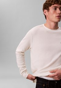 Cremefarvet trøje med lange ærmer og tekstureret, ribbet mønster, rund halsudskæring og tætsiddende manchetter, båret med mørke denimjeans. Calvin Klein-logo synligt.