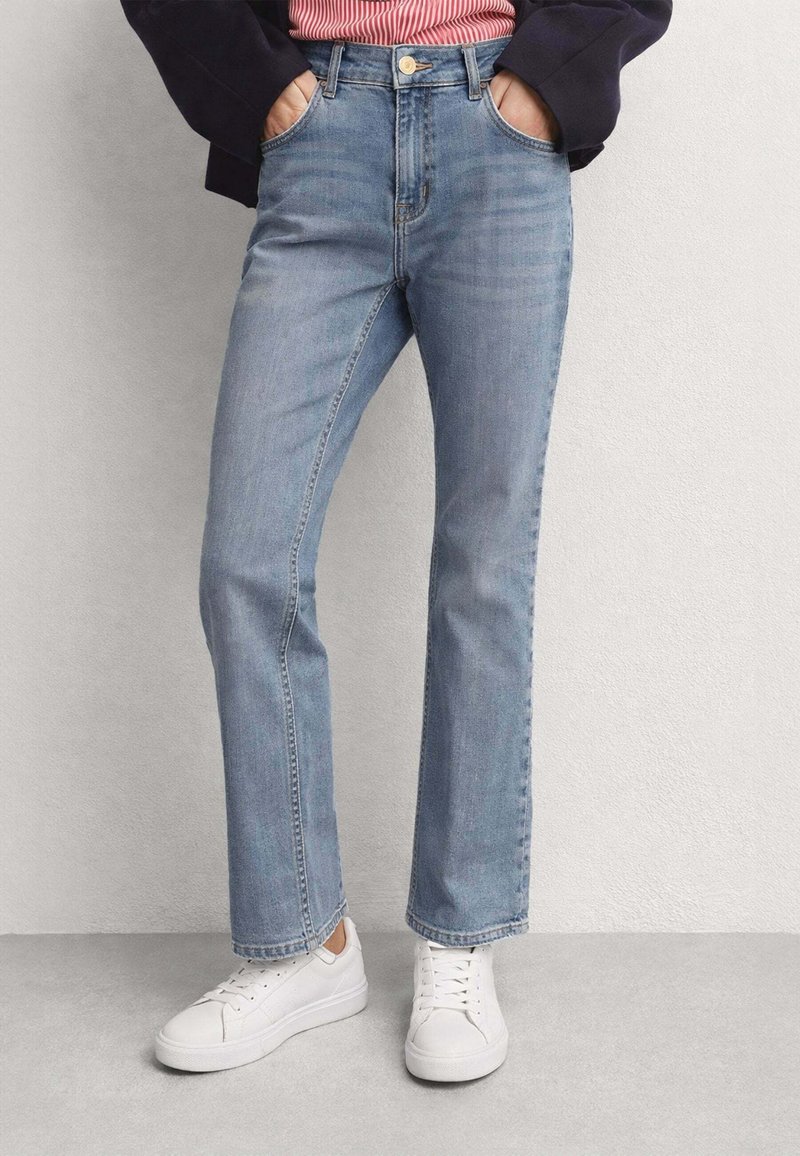 Persona che indossa jeans dritti azzurri con le mani nelle tasche, scarpe da ginnastica bianche e una giacca nera sopra una maglietta a righe rosse.