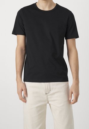 Mężczyzna w prostym czarnym t-shircie z krótkim rękawem i kremowych spodniach stojący na tle jednolitego jasnego tła.