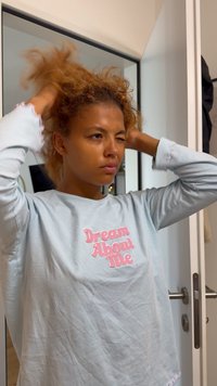 Person som justerar lockigt hår framför en spegel, iklädd en ljusblå skjorta med texten "Dream About Me" i rosa.