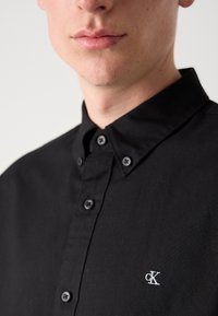 Black button-up shirt, cotton material, featuring a structured collar, front placket with buttons, and a small white "cK" logo on the chest.