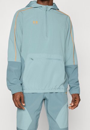 Homme portant une veste à capuche Under Armour bleu clair et gris ainsi qu'un pantalon assorti avec des touches orange, debout devant un fond uni.