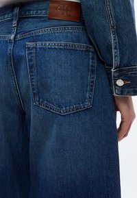 Μπλε τζιν παντελόνι με πίσω τσέπη και καφέ δερμάτινο λογότυπο Calvin Klein, φορεμένο με μανίκι τζιν μπουφάν στο ίδιο χρώμα και ορατό χέρι.