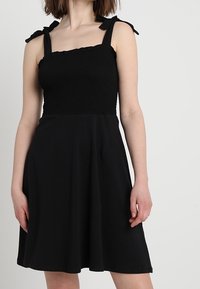 Žena v bezramínkových černých šatech s nařaseným živůtkem a uvázanými ramínky, stojící na pozadí světle jednobarevného pozadí.