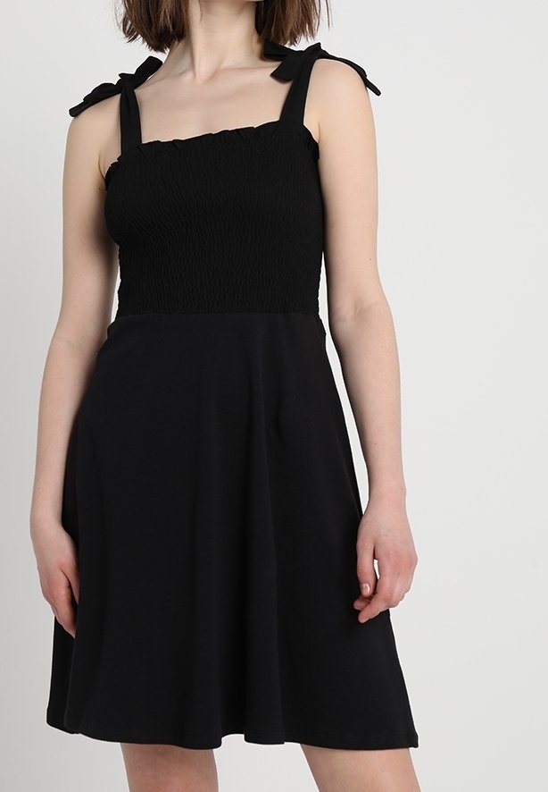 Žena v bezramínkových černých šatech s nařaseným živůtkem a uvázanými ramínky, stojící na pozadí světle jednobarevného pozadí.